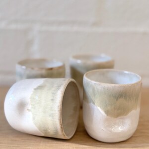 May include: A set of four ceramic cups featuring a two-tone glaze with a creamy white base and a soft sage green drip pattern. Each cup has a smooth, rounded silhouette with a subtle thumb indentation on the side for a comfortable grip.