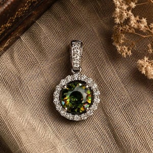 May include: A round pendant featuring a large, faceted olive-green gemstone at the center, surrounded by a halo of small, clear white stones. The metal setting is silver-toned, and the bail is also encrusted with tiny, clear white stones.
