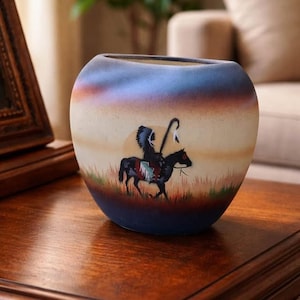 Könnte beinhalten: Eine runde Keramikvase mit einem indianischen Design. Die Vase zeigt eine gemalte Szene eines Indianers auf einem Pferd vor einem Sonnenuntergangshintergrund. Die Vase hat einen blauen Rand und Boden, mit einem cremefarbenen Körper.
