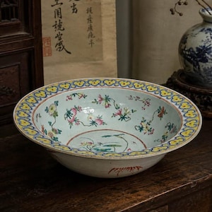 May include: A shallow ceramic bowl with a pale blue interior featuring hand-painted floral and peach motifs. The wide, flared rim is decorated with a repeating yellow and blue cloud-like pattern, while the exterior base displays a red insect design.