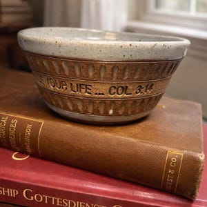 May include: A ceramic bowl with a light blue rim and brown base, inscribed with "YOUR LIFE... COL 3:14", rests on a stack of books. The books have brown and red covers with gold lettering. The scene is set indoors.