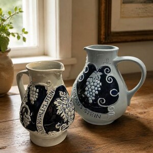 Może przedstawiać: Dwa ceramiczne dzbany z uchwytami. Jeden jest jasnoniebieski z ciemnoniebieskim wzorem winogron i napisem "WEIN AUS STEIN". Drugi jest w kolorze złamanej bieli z ciemnoniebieskim panelem i napisem "AUS KUHLEM STEIN".