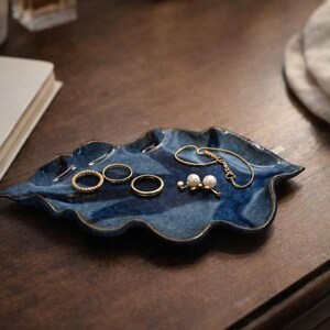 May include: A blue ceramic leaf-shaped jewelry dish with a glossy finish. The dish holds several gold rings, a bracelet, and a pair of pearl earrings. The dish sits on a dark wooden surface.
