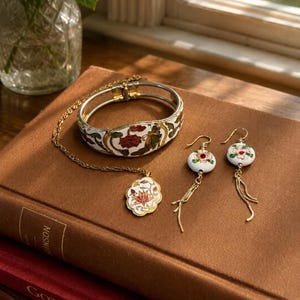 May include: A collection of vintage jewelry featuring a white and gold bracelet, a pendant necklace, and a pair of earrings. The jewelry has a floral design with red and green accents. The pieces are displayed on a brown book.