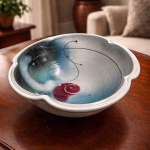 May include: A ceramic bowl with a scalloped edge. The interior is a mix of blue, black, and white, with a red spiral detail. Fine black lines enhance the design. The bowl rests on a dark wooden surface. A decorative item for the home.
