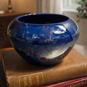 May include: A round, deep blue ceramic bowl with a glossy finish. The bowl sits atop two antique books with gold lettering. The bowl's surface reflects the surrounding environment, creating a unique visual effect. The books are brown and red.