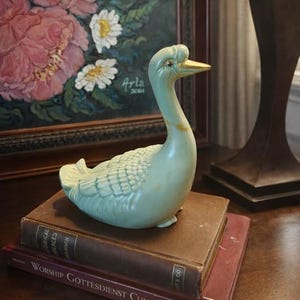 Puede incluir: Una figura de cisne de cerámica verde claro con pico dorado, sobre una pila de libros antiguos. El cisne tiene detalles de plumas texturizadas. Una pintura de flores rosas en el fondo.