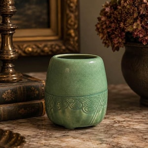 May include: A cylindrical ceramic vessel in a matte sage green finish. The upper portion is smooth, while the lower section features a raised, embossed pattern of swirling lines and vertical ridges. The base has small, subtle feet for stability.