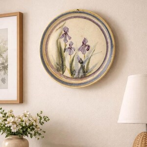Puede incluir: Un plato decorativo redondo con un diseño floral pintado a mano con iris moradas. El plato tiene un centro color crema con un borde azul y amarillo. Está colgado en una pared color crema, añadiendo un toque artístico al espacio.