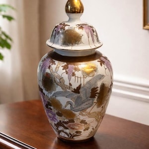 May include: A decorative ceramic jar with a lid, featuring a gold finial. The jar is white with a painted design of cranes, wisteria, and gold accents. The jar is sitting on a wooden surface.