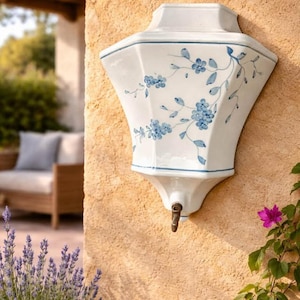 Puede incluir: Fuente de agua de cerámica blanca para pared con diseño floral azul. Cuenta con un grifo de latón y está montada en una pared texturizada de color marrón claro. Elemento decorativo para jardín exterior.