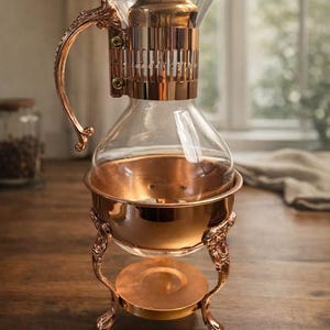 Puede incluir: Una cafetera de sifón de color cobre con una jarra de vidrio transparente y un asa decorativa. La cafetera se encuentra sobre un soporte de color cobre con patas ornamentadas y una base redonda. El diseño es vintage y elegante.