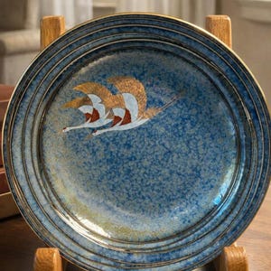 Könnte beinhalten: Eine blaue Keramikplatte mit goldenem Rand und einem Design von fliegenden Vögeln. Die Platte wird auf einem Holzständer präsentiert, der ihren dekorativen Charakter hervorhebt. Die Oberfläche der Platte hat eine gesprenkelte Textur.
