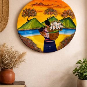 Può includere: Opera d'arte murale rotonda con una vivace pittura di paesaggio. L'opera raffigura una persona che trasporta un cesto di fiori bianchi, sullo sfondo di montagne verdi, un lago blu e un tramonto arancione. Arte murale decorativa.