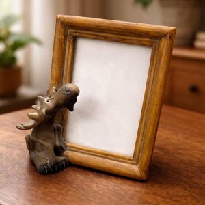 Peut inclure: Un cadre photo en bois avec une finition marron clair, posé sur une surface en bois. Une petite figurine d'orignal est positionnée à gauche du cadre. Le cadre a un intérieur blanc, prêt pour une photo.