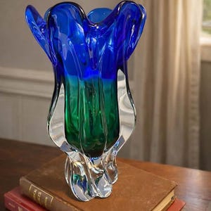 Op de afbeelding: Een decoratieve glazen vaas met een uitlopende, organische vorm. De vaas heeft een kleurovergang, van diepblauw aan de bovenkant naar groen in het midden, met een heldere basis. Hij staat op een stapel vintage boeken.