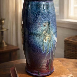 May include: A tall, ceramic vase with a glossy finish. The vase transitions from deep purple at the base to shades of blue and teal towards the top, with a dark rim. It sits on a stack of books.