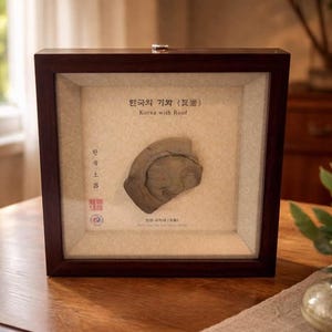 May include: A framed Korean artifact, possibly a roof tile fragment, displayed in a dark wooden frame. The artifact is a light brown color and has a detailed relief design. The frame includes text in Korean and English, with the title "Korea with Roof".