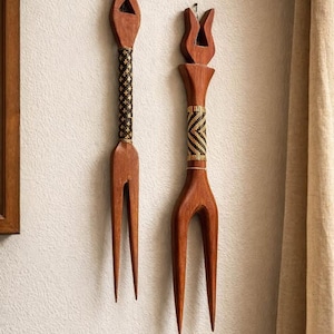 Puede incluir: Dos tenedores decorativos de madera con una parte superior en forma de tulipán están montados en una pared texturizada. Los mangos presentan detalles tejidos en negro y dorado. Los tenedores son de color marrón cálido y añaden un toque rústico a la decoración.