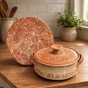 Puede incluir: Una fuente de cerámica y un plato a juego. El plato presenta un diseño en relieve detallado de hojas y espirales. La fuente tiene tapa y asas, con un patrón en relieve similar. El color es naranja y crema cálidos y terrosos.