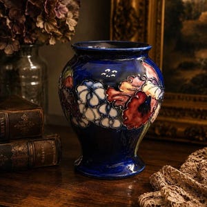 May include: A deep blue ceramic vase featuring a raised, hand-painted floral motif with white, red, and green petals. The vase has a rounded body, a flared rim, and a glossy finish, showcasing intricate relief work on the exterior surface.
