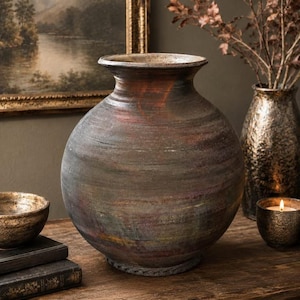 May include: A large, spherical ceramic vase with a wide, flared rim. The surface features a textured, weathered finish in muted tones of charcoal gray, deep brown, and subtle hints of iridescent copper and bronze throughout the rounded body.