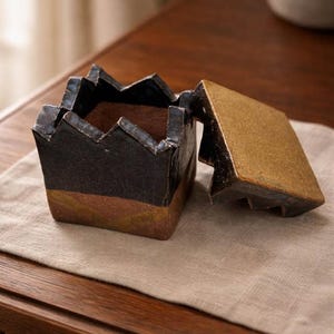 Może przedstawiać: Ręcznie wykonane ceramiczne pudełko z pokrywką, o unikalnym wzorze przypominającym koronę. Pudełko jest głównie czarne z brązowo-złotą podstawą. Pokrywka jest otwarta, odsłaniając wnętrze. Pudełko leży na lnianej tkaninie w neutralnym kolorze.