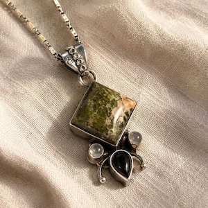 Könnte beinhalten: Eine Halskette mit Anhänger aus Sterlingsilber mit einem quadratisch geschliffenen, grün-rosa gesprenkelten Stein, zwei kleinen runden weißen Steinen und einem birnenförmigen schwarzen Stein. Die Silberöse ist mit 925 gestempelt und an einer flachen Gliederkette befestigt.
