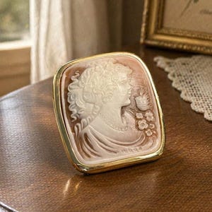 May include: A vintage cameo brooch with a gold-tone frame. The cameo features a detailed profile of a woman with flowing hair and a floral accent. The brooch is rectangular with rounded corners, showcasing a classic, elegant design.