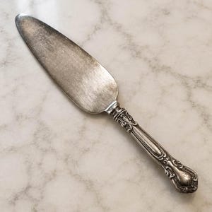 Puede incluir: Un servidor de pasteles de tono plateado con una hoja ancha, plana y cónica que presenta rayones superficiales visibles y un mango decorativo adornado con intrincados relieves y detalles verticales a lo largo de la empuñadura.