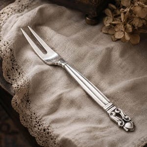 May include: A silver-toned metal serving fork featuring two long, slender tines and a decorative handle with an ornate, raised scrollwork pattern at the base. The utensil has a polished finish with subtle darkened accents in the recessed areas.