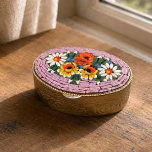 Puede incluir: Una caja de baratijas ovalada con un diseño floral de mosaico. La tapa presenta un mosaico de flores rosas, blancas, amarillas y naranjas. La caja tiene una base de metal dorado con detalles en relieve y un borde con cuentas.