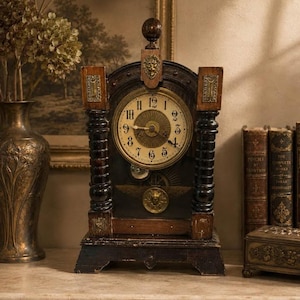 Puede incluir: Un reloj antiguo de madera con esfera redonda, números romanos y detalles ornamentados. El reloj tiene un acabado marrón oscuro con detalles dorados y está flanqueado por pilares decorativos. El reloj se exhibe sobre una superficie de mármol, con libros y un jarrón al fondo.