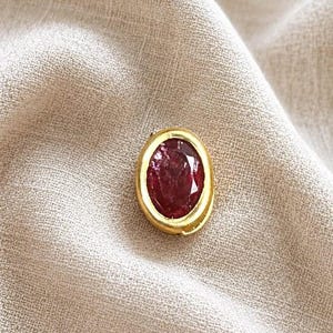 May include: A gold-colored, oval-shaped pendant with a large, deep red gemstone. The gemstone is set within a gold bezel, and the pendant is resting on a beige fabric surface. The gemstone has a faceted appearance.