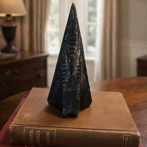 Op de afbeelding: Een zwarte, piramidevormige sculptuur met getextureerde details, rustend op een stapel boeken. De sculptuur heeft een ruwe, verweerde uitstraling. De boeken zijn gebonden in bruine en rode covers, met gouden letters. De scène speelt zich af op een houten tafel.