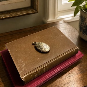 May include: An antique silver locket with an ornate floral design rests on a brown book. The book is stacked on top of a red book. The scene is set on a wooden table, with a window and a potted plant in the background.