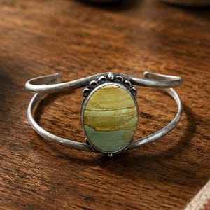 May include: A silver cuff bracelet featuring a central oval-shaped stone with horizontal bands of yellow, tan, and pale green. The metal band has a polished finish with a decorative beaded border surrounding the stone setting at the center.