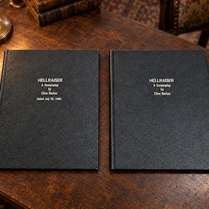 Könnte beinhalten: Zwei identische schwarze Hardcover-Bücher mit strukturierter Oberfläche. Auf den Vorderseiten steht in weißer Schrift: HELLRAISER, A Screenplay, by, Clive Barker, Dated July 28, 1986. Die Bücher liegen nebeneinander auf einer dunklen Holzoberfläche.