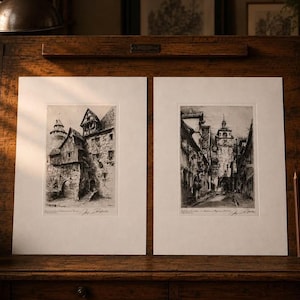 May include: Two black and white architectural etchings on white paper mounts. The left print depicts a stone building with a tower, while the right print shows a narrow street leading to a clock tower. Both prints feature handwritten signatures below.