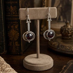 May include: A pair of silver-toned drop earrings featuring a circular dark red gemstone set within a metallic frame. The earrings have a vertical bar design and are suspended from silver hooks, displayed on a beige T-shaped jewelry stand.