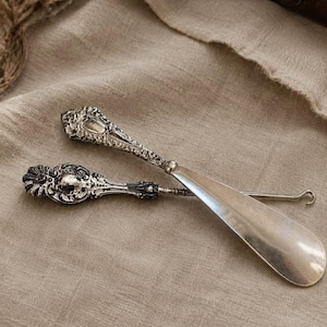 May include: A silver-toned metal shoehorn and a button hook, both featuring ornate, embossed handles with intricate scrollwork and floral detailing. The shoehorn has a smooth, curved blade, while the button hook ends in a small, thin metal loop.