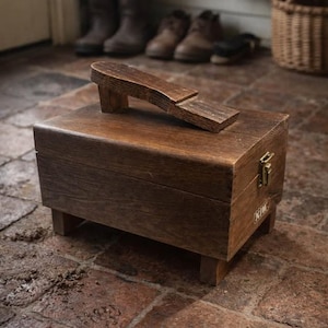 Könnte beinhalten: Eine Holzschuhputzkiste mit eingebauter Schuhablage. Die Kiste ist rechteckig mit einem Klappdeckel und steht auf kurzen Beinen. Die Schuhablage ist abgewinkelt und oben befestigt. Die Kiste ist aus dunkelbraunem Holz.
