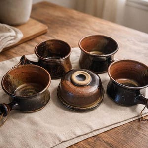 Puede incluir: Un conjunto de cuencos de cerámica hechos a mano y una tapa. Los cuencos son de color marrón oscuro con interiores y asas de color marrón claro. La tapa es redonda con una abertura central. Los artículos están dispuestos sobre un paño de lino beige.
