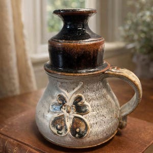 Puede incluir: Una taza de cerámica hecha a mano con un diseño único. La taza presenta un cuerpo moteado gris claro y marrón, un asa y un trébol decorativo. La parte superior es de color marrón oscuro y negro. La taza está colocada sobre una pila de libros.