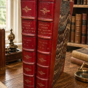 May include: Two antique red leather-bound books titled England Under the House of Hanover by Wright, Volume I and Volume II. The spines feature gold embossed lettering and decorative floral motifs, with marbled paper covers on the side panels.
