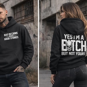 Könnte beinhalten: Schwarzer Kapuzenpullover mit weißem Text: "BAD DECISIONS MAKE GOOD STORIES" auf der Vorderseite. Ein weiterer schwarzer Hoodie mit dem Text "YES I'M A BITCH BUT NOT YOURS" und einer Totenkopf-Grafik auf dem Rücken.