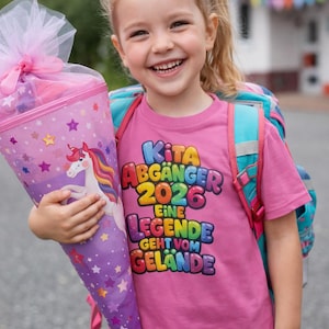 May include: A smiling young girl wearing a pink t-shirt with the text "KITA ABGÄNGER 2026 EINE LEGENDE GEHT VOM GELÄNDE" and a backpack, holding a purple and pink school cone decorated with stars and a unicorn.