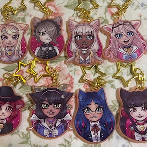 Op de afbeelding: Acht acryl sleutelhangers met anime-achtige karakters met kattenoren. Elke sleutelhanger heeft een goudkleurige ketting en een stervormige bedel. De karakters hebben verschillende haarkleuren en outfits, tegen een roze achtergrond.