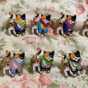 Op de afbeelding: Een verzameling emaille pins met een cartoon schildpadkat ontwerp. Elke pin heeft een gouden omtrek en toont een kat die een hoodie draagt in verschillende pride vlag kleuren, waaronder regenboog, roze, blauw en groen. De pins zijn gerangschikt op een bloemenstof.