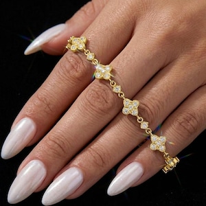 May include: A gold-colored bracelet featuring a clover design with clear, sparkling stones. The bracelet is composed of multiple clover-shaped links connected by smaller, round stones. The bracelet is displayed on a hand with long, white fingernails.
